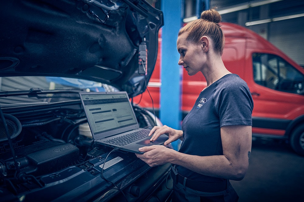 Technik używa laptopa do diagnozowania furgonetki Ford Transit w warsztacie, z inną czerwoną furgonetką w tle.