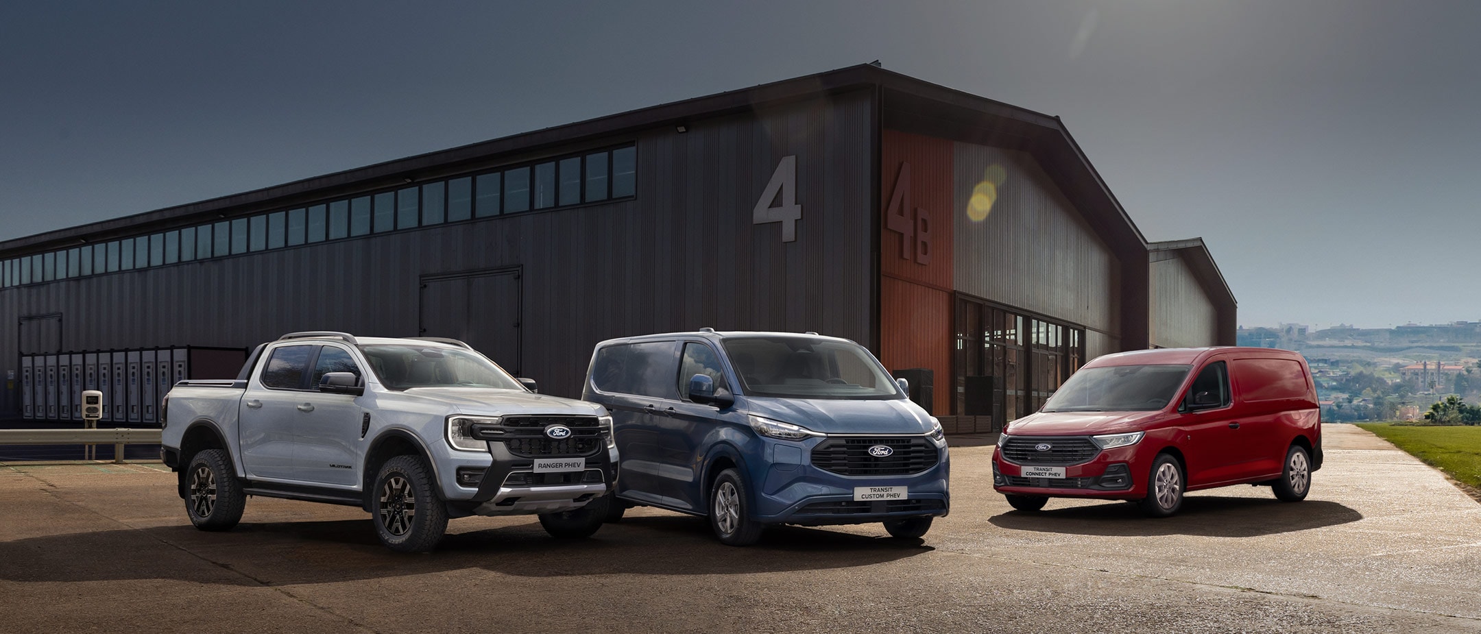 Trzy samochody marki Ford – srebrny pick-up oraz niebieski i czerwony furgon – zaparkowane przed dużym magazynem w słoneczny dzień.