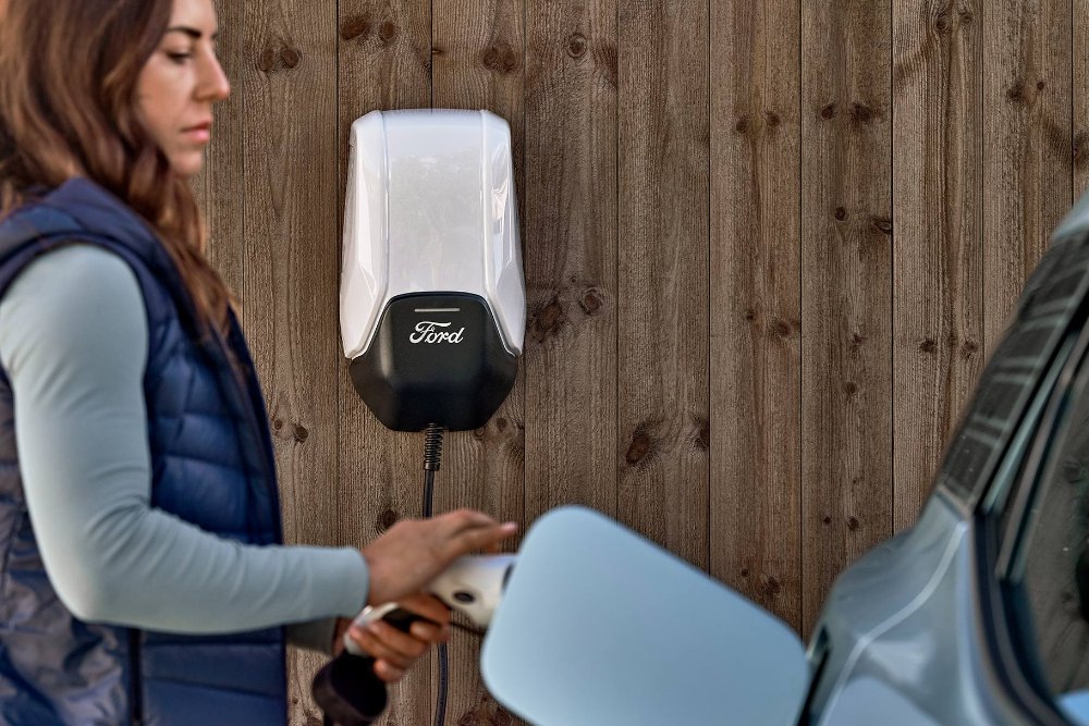 Kobieta podłącza niebieskiego elektrycznego Forda Explorera do domowej stacji ładowania pojazdów elektrycznych.