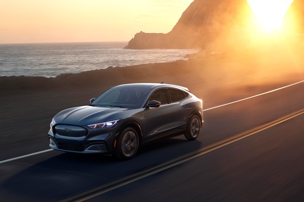 Elektryczny Ford Mustang Mach-E jedzie nadmorską drogą o zachodzie słońca.