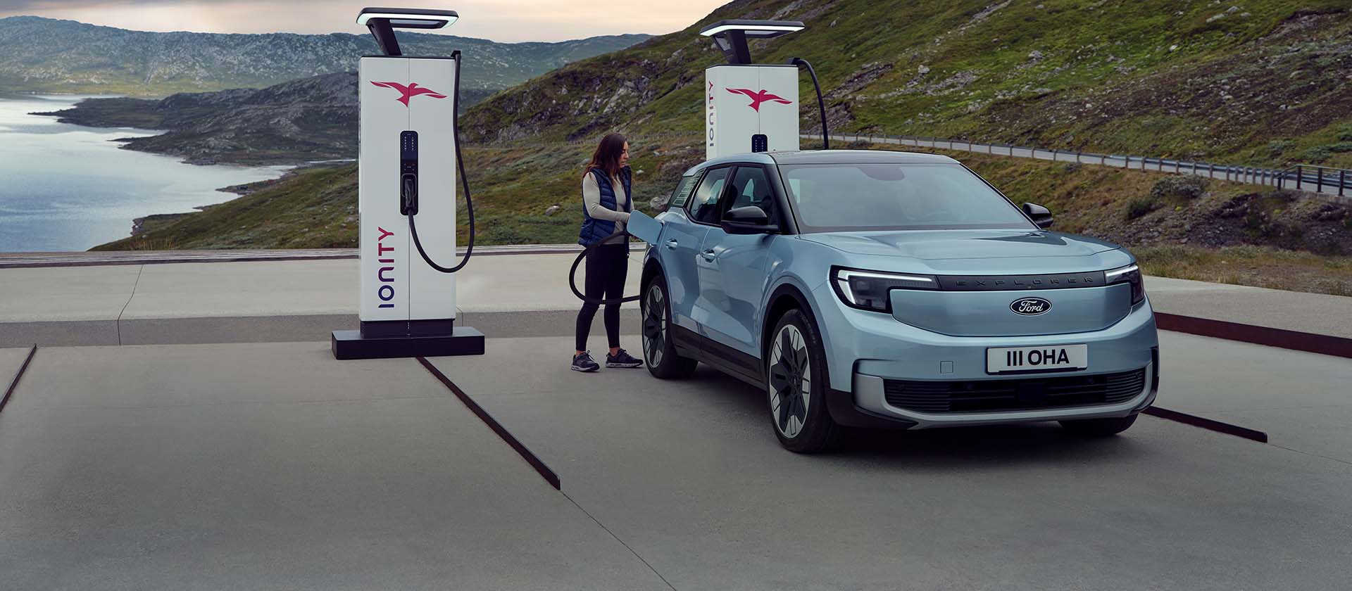 Kobieta podłącza niebieskiego elektrycznego Forda Explorer do publicznej stacji ładowania za pomocą kabla. Samochód jest zaparkowany w pobliżu gór i wody. 