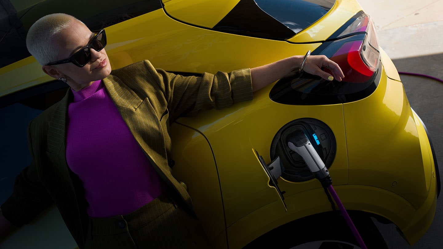 Kobieta opierająca się o żółtego elektrycznego Forda Capri, podłączonego do ładowarki.