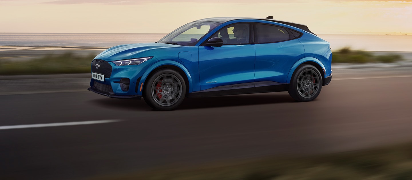 Niebieski elektryczny Ford Mustang Mach-E jadący nadmorską drogą o zachodzie słońca.