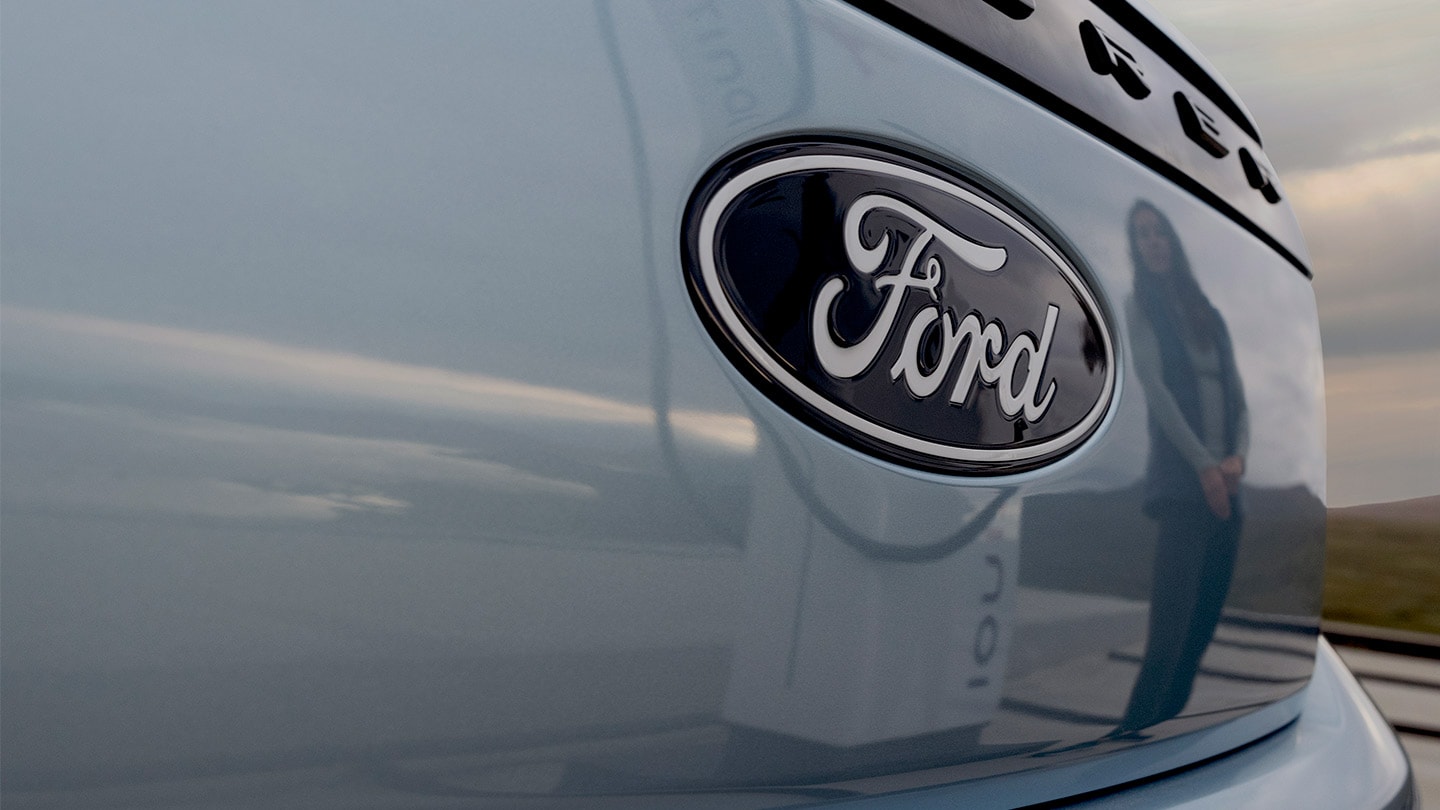 Logo Forda na niebieskim elektrycznym Fordzie Explorer, z odbiciem publicznej stacji ładowania w samochodzie.