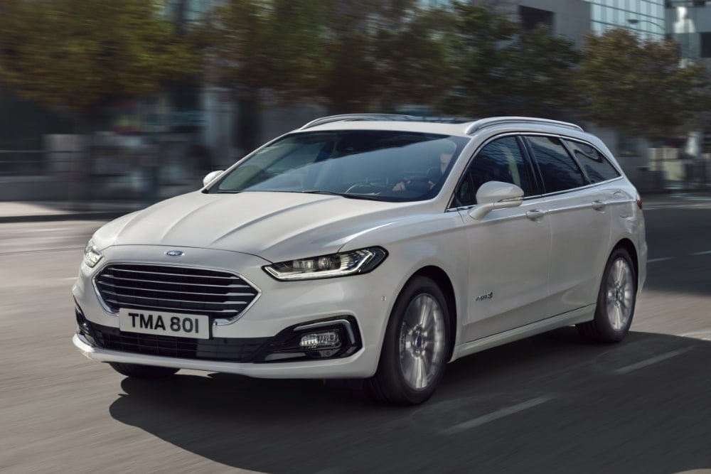 Biały Ford Mondeo Hybrid kombi jedzie ulicą miasta, uchwycony z przedniego trzy czwarte.