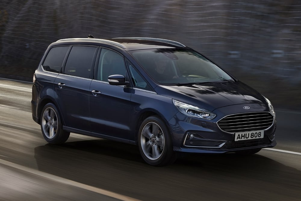 Granatowy Ford Galaxy jedzie drogą o zmierzchu, uchwycony z przedniego trzy czwarte.