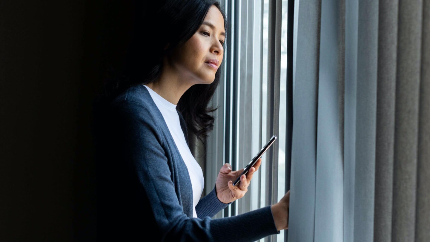 A woman holding a smartphone looking outside the window