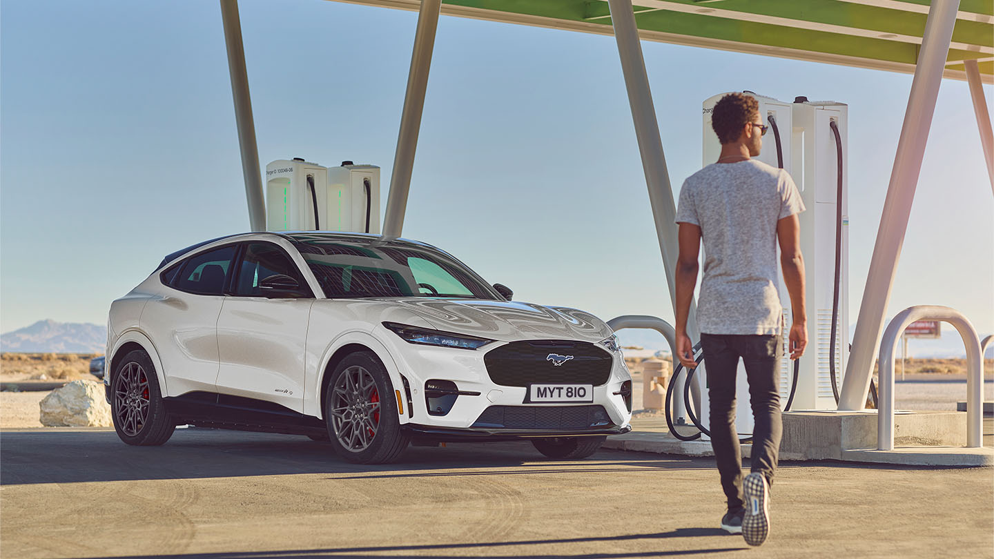 Biały Ford Mustang Mach-E zaparkowany przy publicznej stacji ładowania. Mężczyzna w białej koszulce idzie w stronę samochodu.