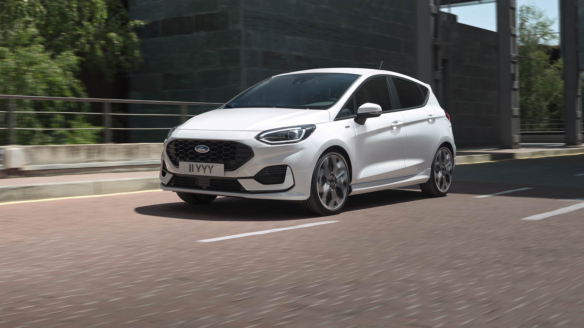 Biały Ford Fiesta poruszający się po mieście z zielenią i budynkami w tle.