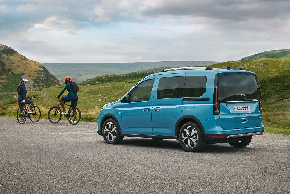 Niebieski Ford Tourneo Connect zaparkowany na drodze w zielonym, pagórkowatym krajobrazie, z dwoma rowerzystami w pobliżu.