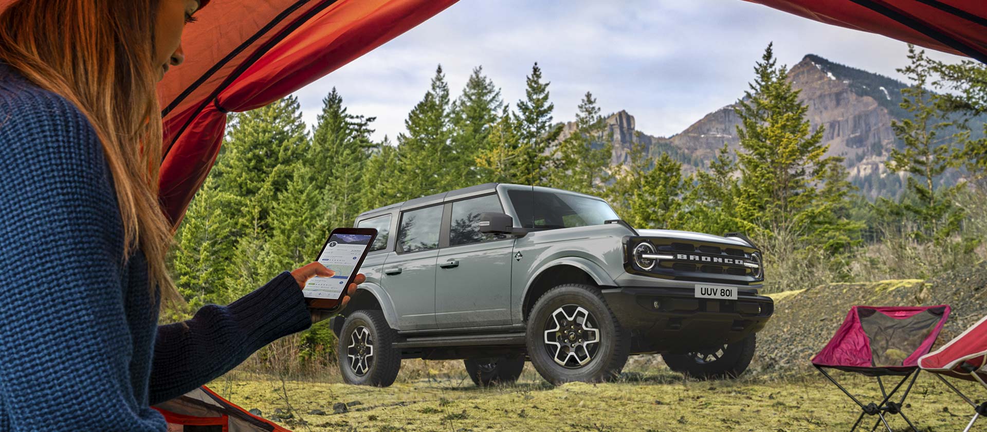 Widok Forda Bronco w tle na kempingu górskim, z osobą z telefonem w namiocie na pierwszym planie.