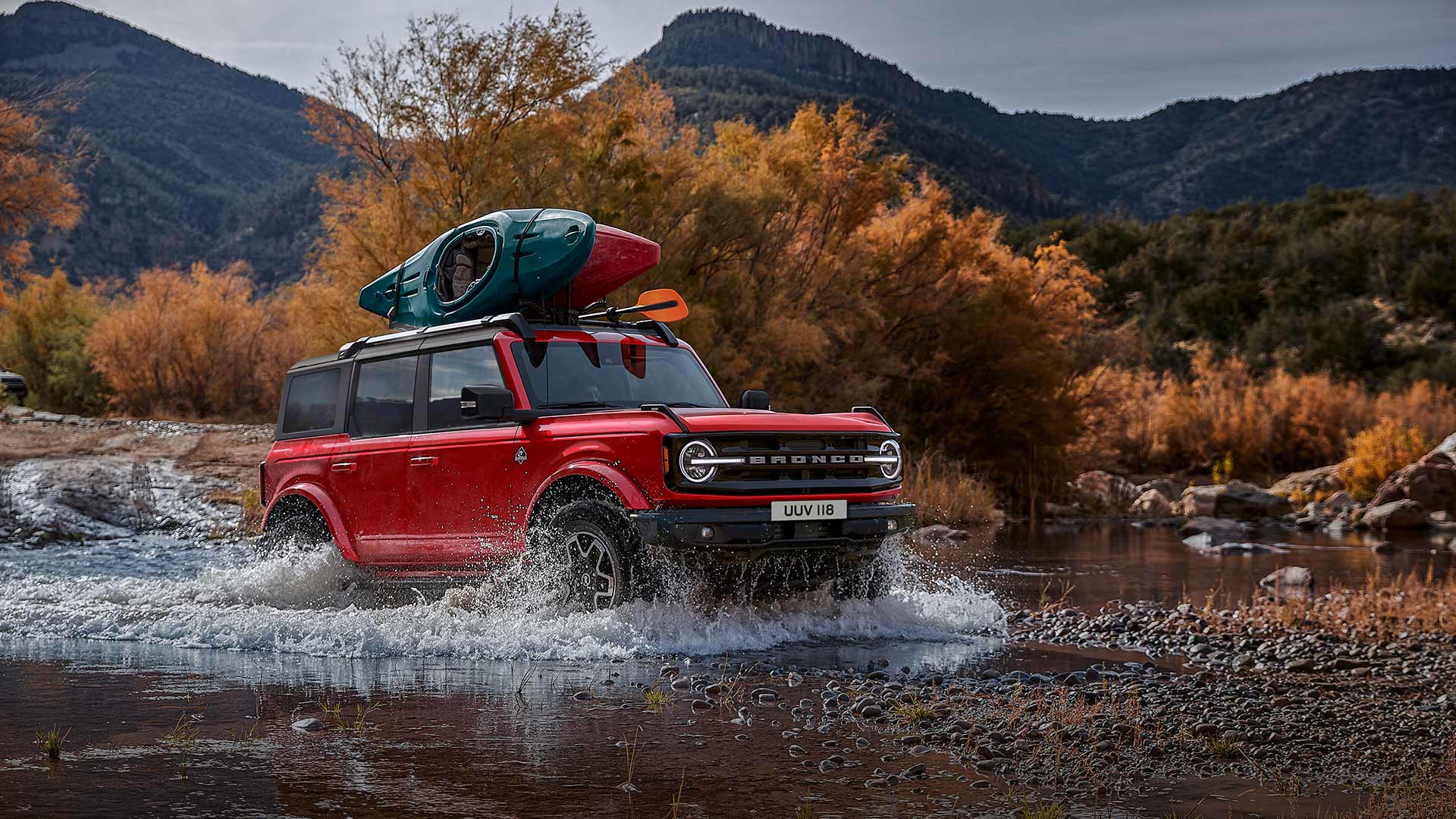 Widok z boku czerwonego Forda Bronco przejeżdżającego przez płytką wodę w górach.