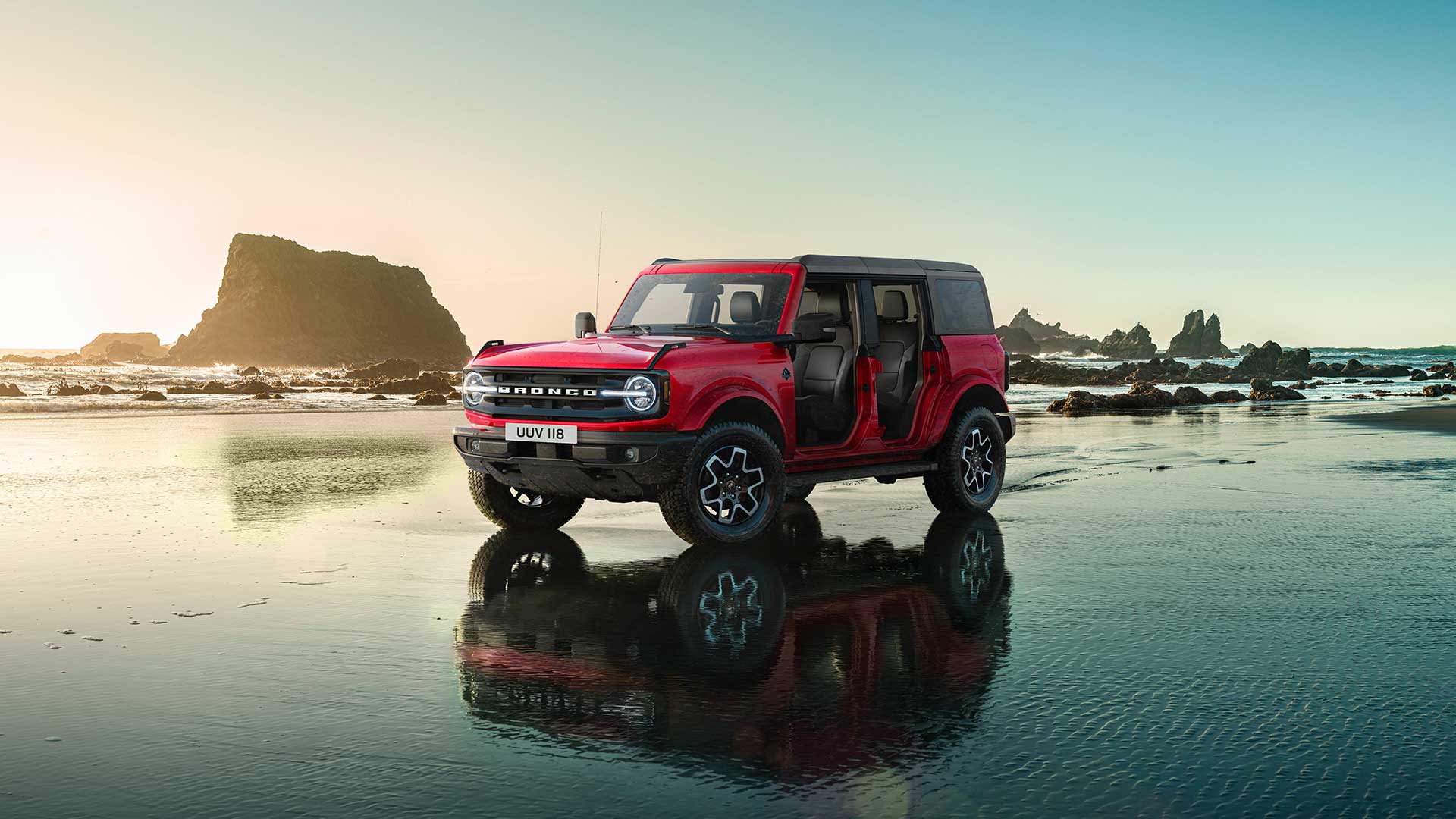 Widok z boku czerwonego Forda Bronco, ze zdjętymi drzwiami, zaparkowanego na mokrym, piaszczystym brzegu.