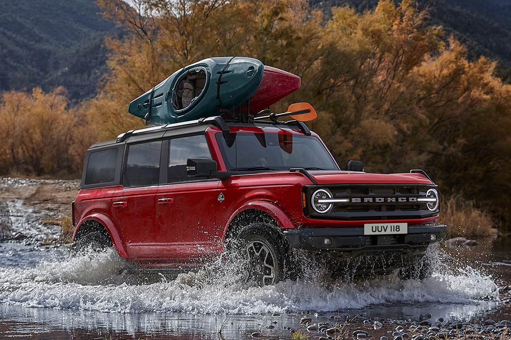 Widok z boku czerwonego Forda Bronco przejeżdżającego przez płytką wodę w górach.