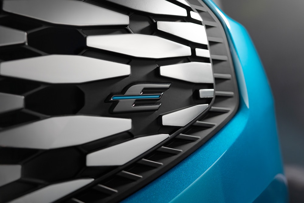 Zbliżenie na osłonę chłodnicy Forda E-Tourneo Custom z niebieskim logo „E”, zarezerwowanym dla pojazdów elektrycznych marki Ford.