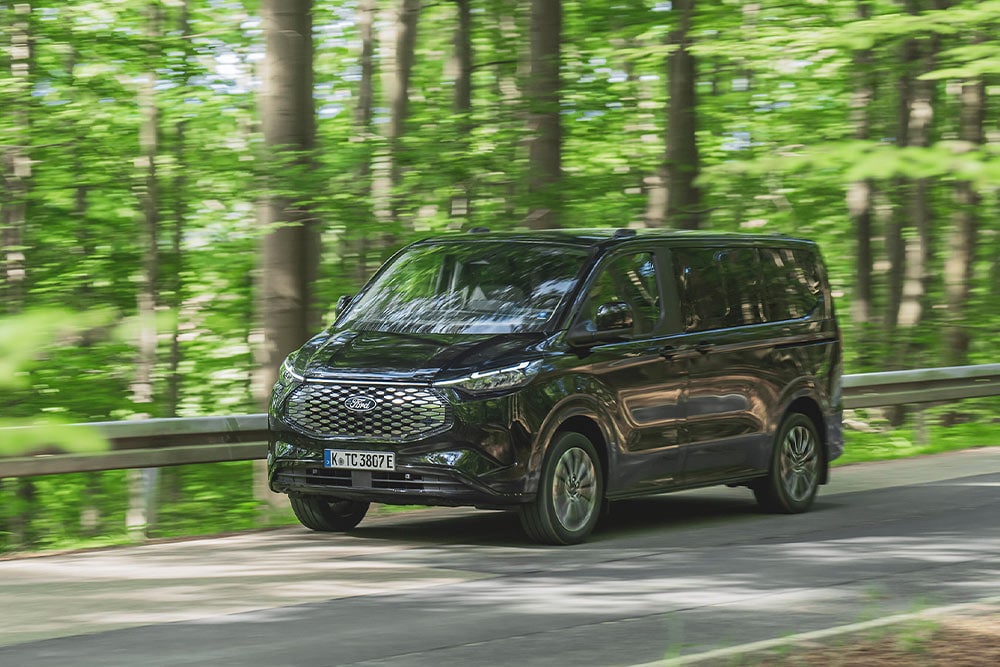 Czarny Ford E-Tourneo Custom jedzie drogą wysadzaną drzewami.