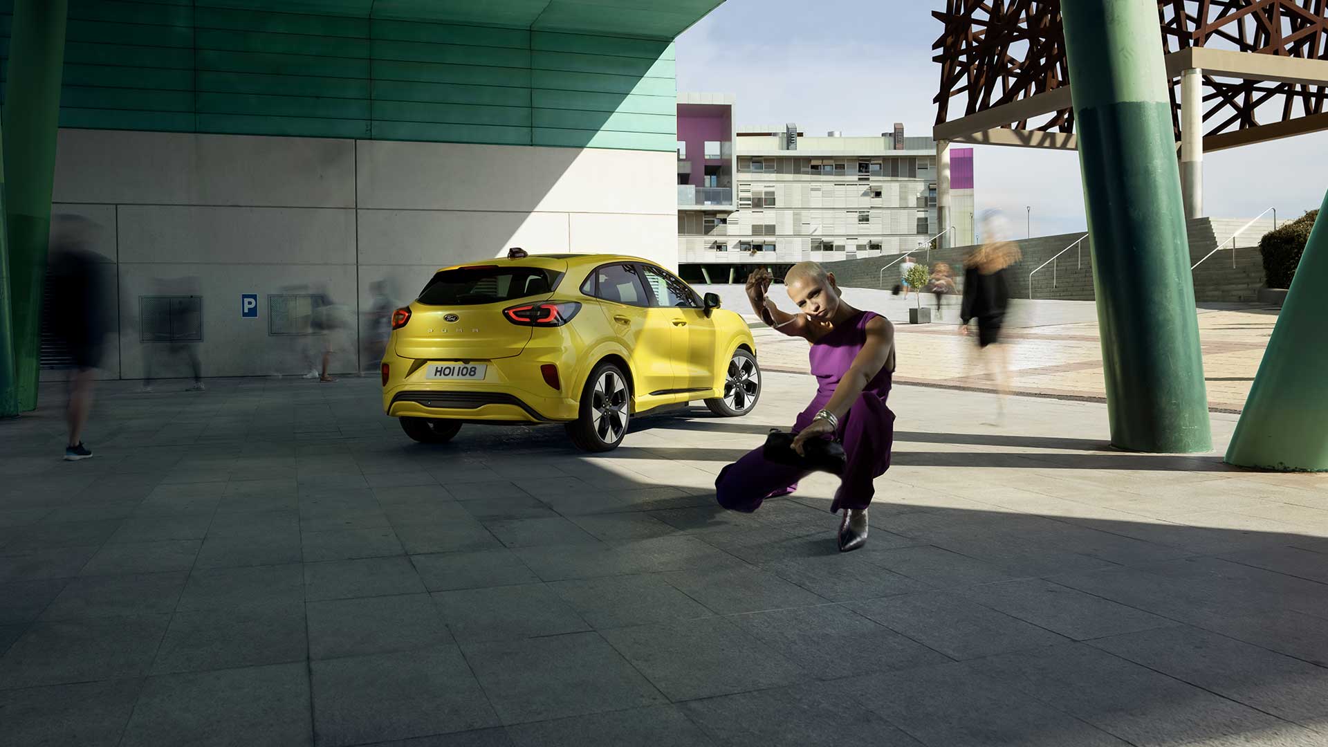 Widok z tyłu na żółtego elektrycznego Forda Puma Gen-E® zaparkowanego pod mostem, obok osoby ubranej na fioletowo.