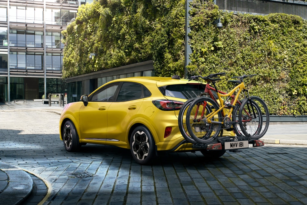 Żółty Ford Puma Gen-E zaparkowany na miejskim parkingu z dwoma rowerami zamontowanymi na tylnym bagażniku.