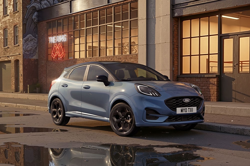 Niebieski Ford Puma zaparkowany przy ulicy o zachodzie słońca. W kałużach widoczne są odbicia otaczających budynków.