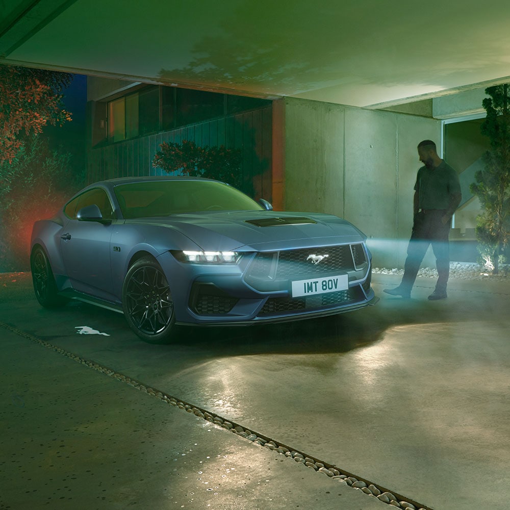 Mężczyzna podchodzący do zaparkowanego Ford Mustang z włączonymi reflektorami, pokazujący powitalne oświetlenie wykrywania podejścia