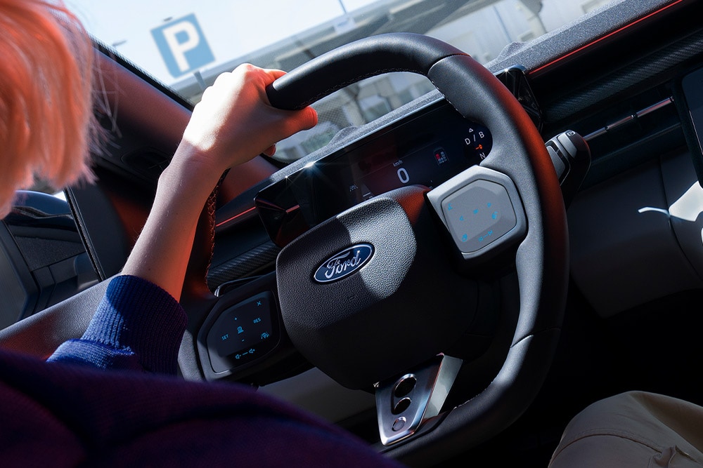 Zbliżenie na kobietę trzymającą kierownicę w zaparkowanym Fordzie Capri, pokazujące również cyfrową tablicę rozdzielczą.