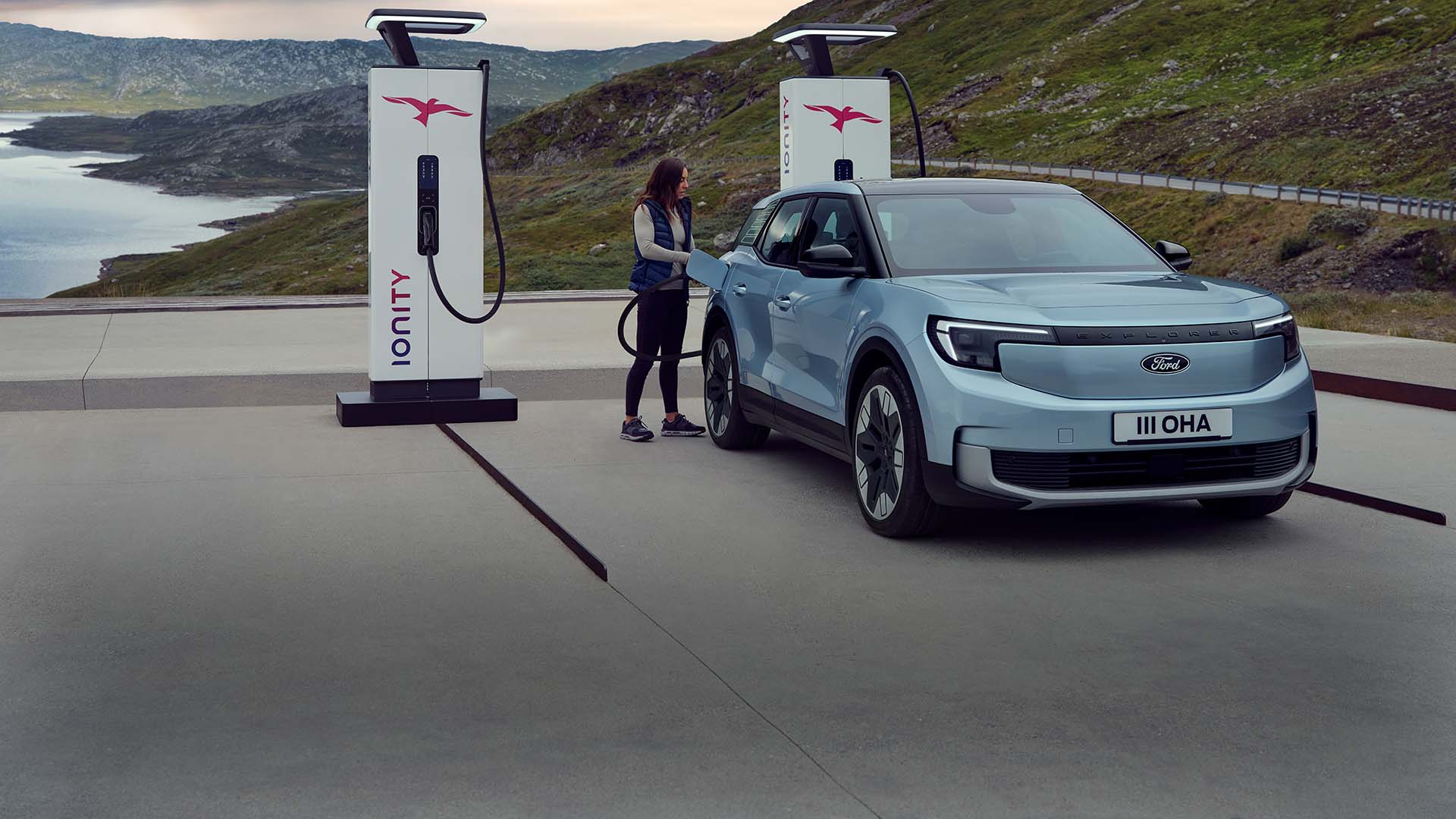 A woman plugs a blue Ford Explorer EV into a public charging station using a cable. The car is parked by mountains and water.