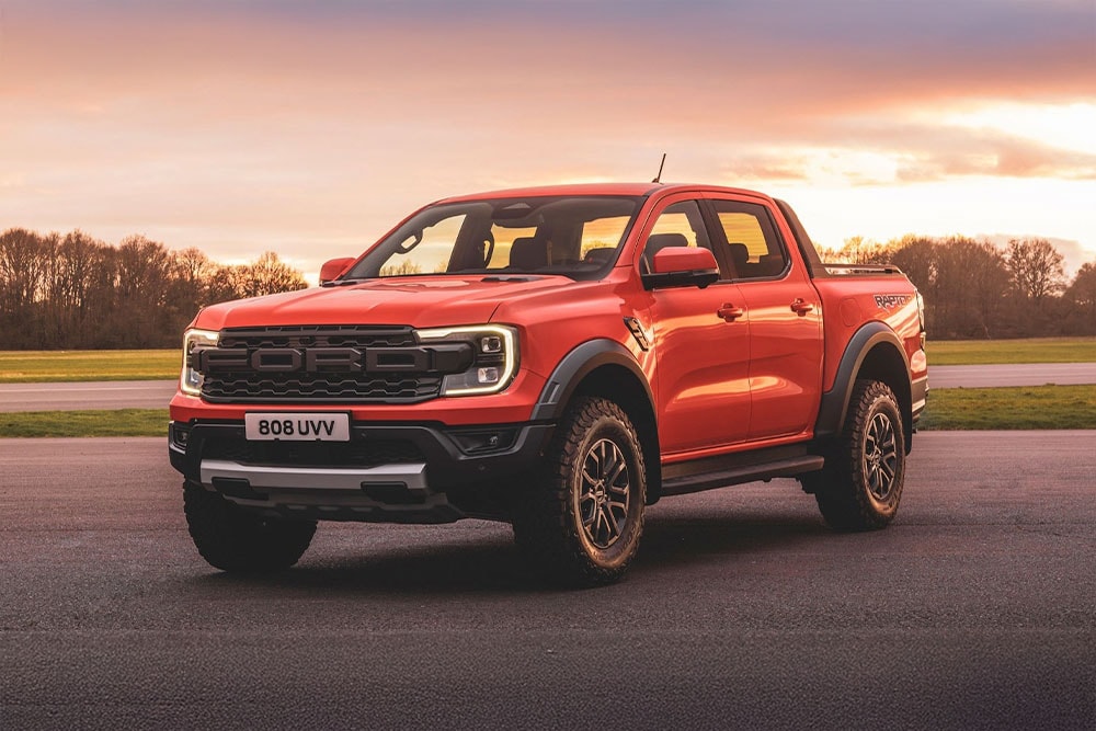 Pomarańczowy pick-up Ford Ranger Raptor zaparkowany na tle malowniczego krajobrazu.