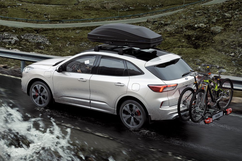 Srebrny Ford Kuga jedzie górską drogą z boxem dachowym i bagażnikiem rowerowym z tyłu.