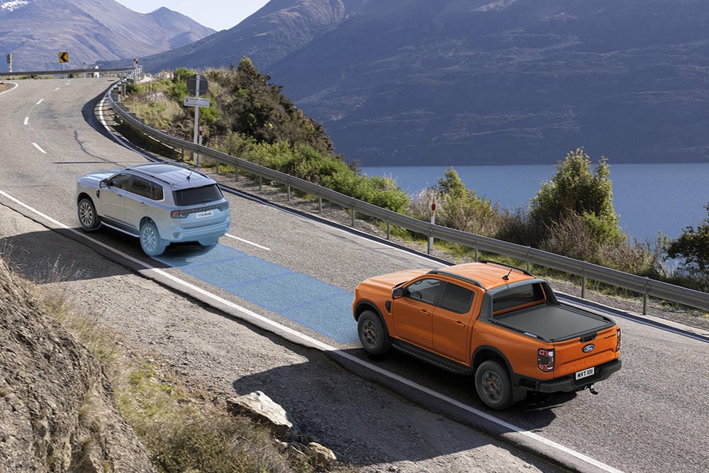 Żółty Ford Ranger Raptor jadący malowniczą górską drogą z widocznymi systemami wspomagającymi kierowcę.