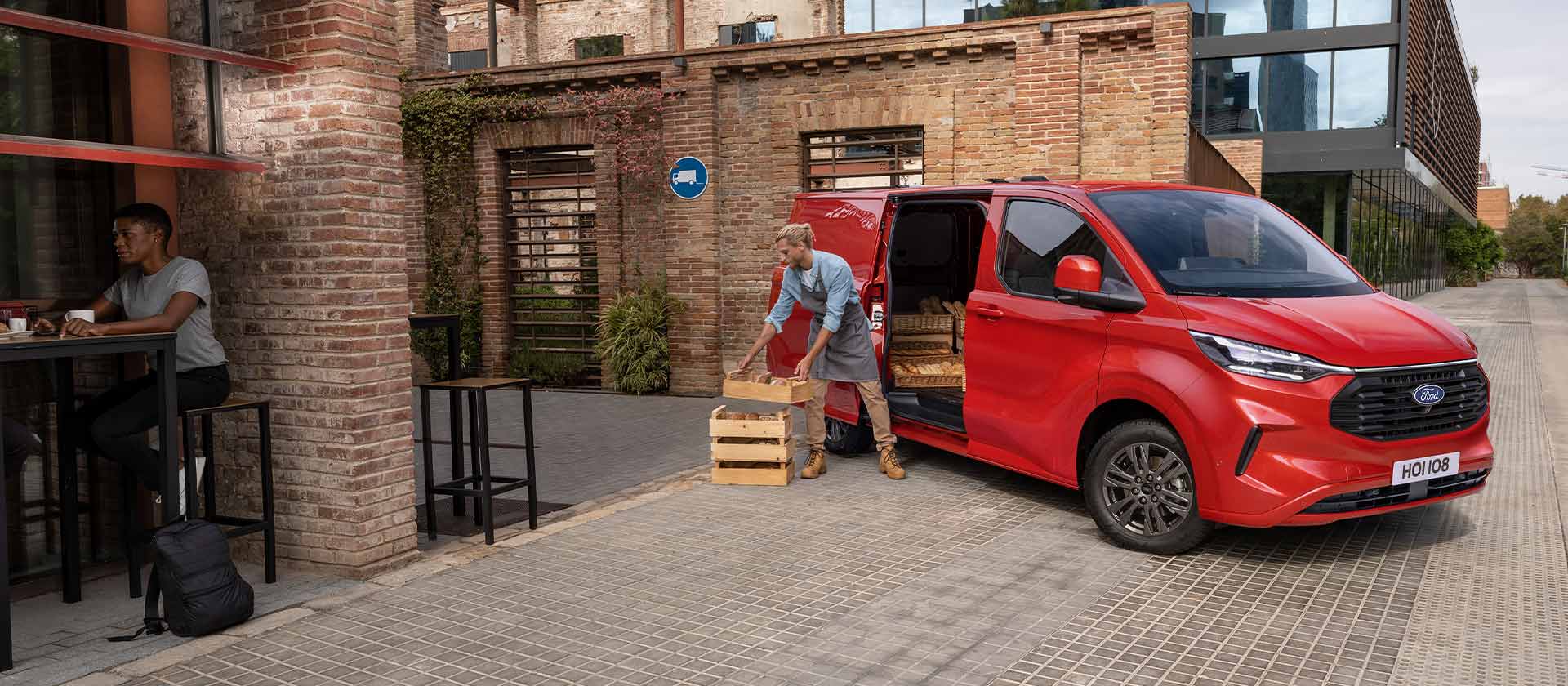 Osoba rozładowuje skrzynie z czerwonego vana Ford Transit Custom zaparkowanego przed kawiarnią. 