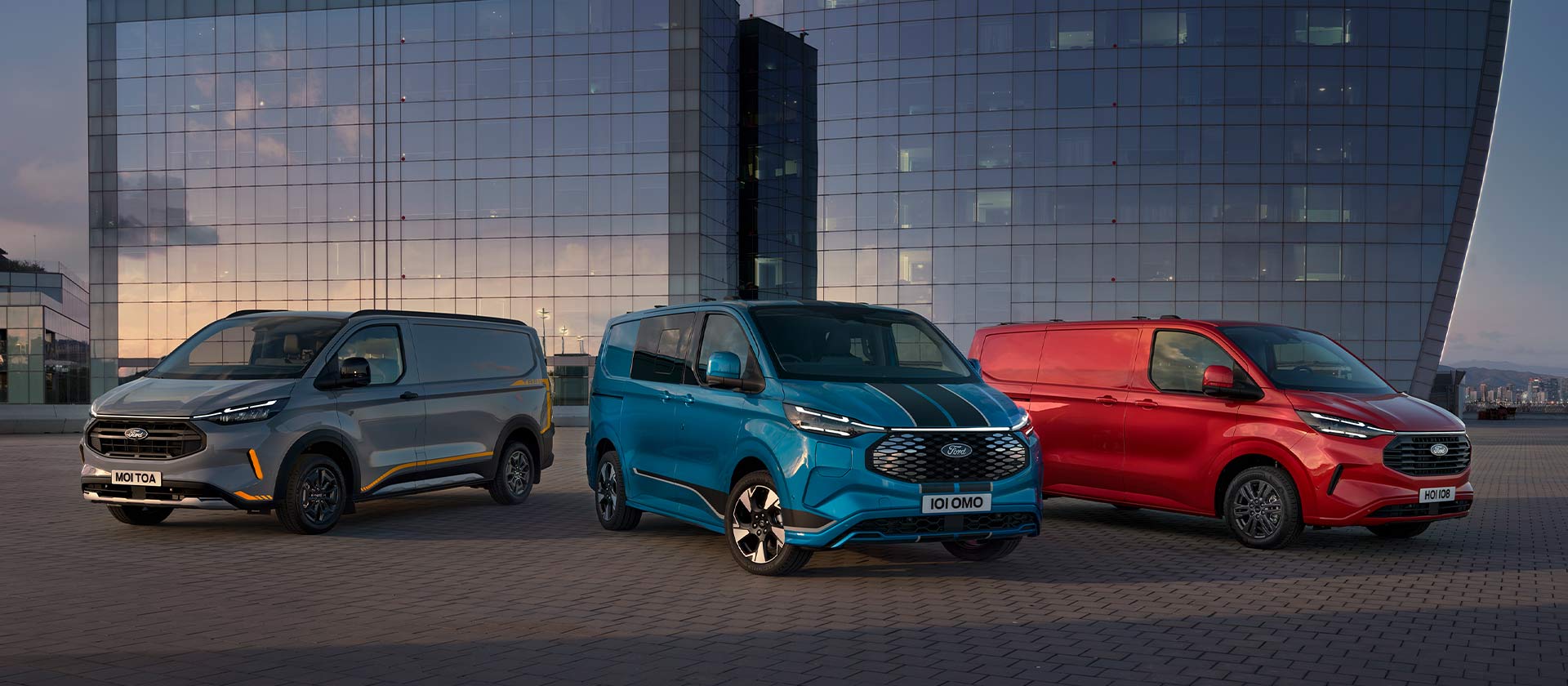 Trzy zaparkowane vany Ford Transit Custom - Trail w kolorze szarym, brygadowy Sport w kolorze niebieskim oraz Limited w kolorze czerwonym. 