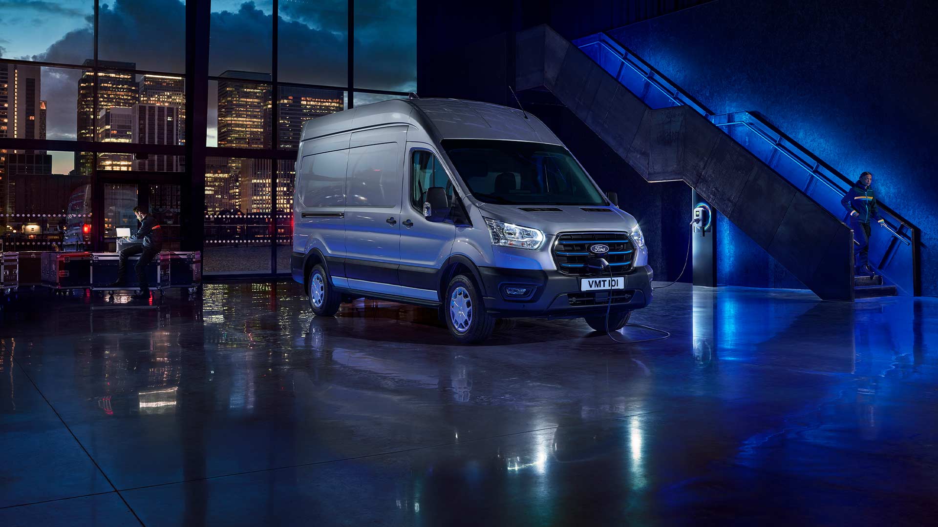 Srebrny elektryczny van Ford E-Transit zaparkowany w pomieszczeniu z panoramą miasta widoczną przez duże okna.