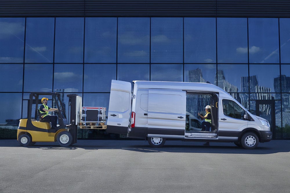 Dwie osoby umieszczające ładunek w srebrnym vanie Ford Transit za pomocą wózka widłowego.