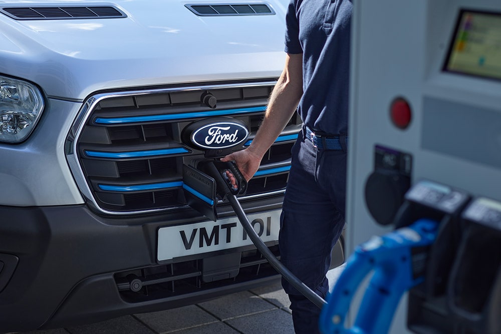 Zbliżenie na osobę podłączającą kabel ładujący do srebrnego elektrycznego vana Ford E-Transit.