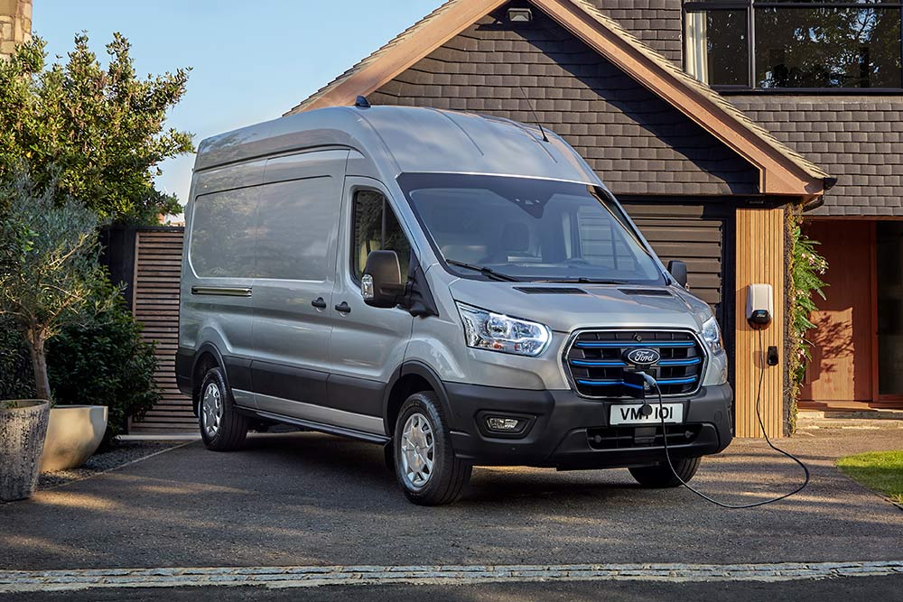 Srebrny Ford E-Transit zaparkowany na podjeździe obok domu i podłączony do stacji ładowania.