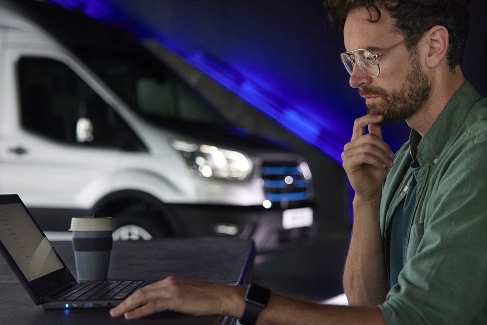 Mężczyzna pracujący na laptopie oraz elektryczny van Ford E-Transit w tle.