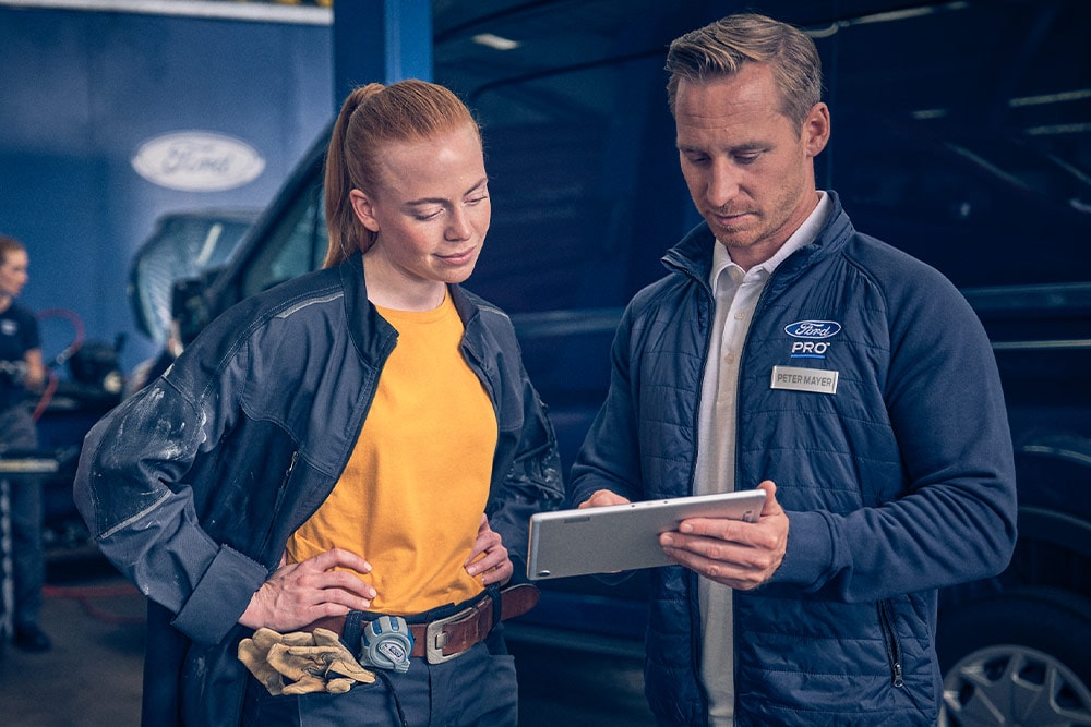 Dwóch mechaników w warsztacie patrzy na tablet przed elektrycznym vanem Ford E-Transit.