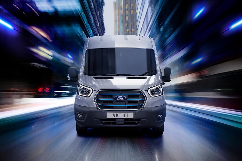 Szary Ford Transit z niebieskimi akcentami na osłonie chłodnicy jedzie nocą przez miasto.