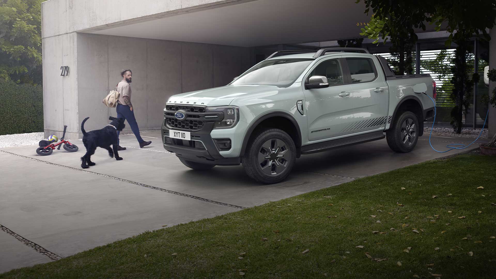 Mężczyzna i pies idą w kierunku szarego hybrydowego Forda Ranger PHEV ładującego się na podjeździe. 