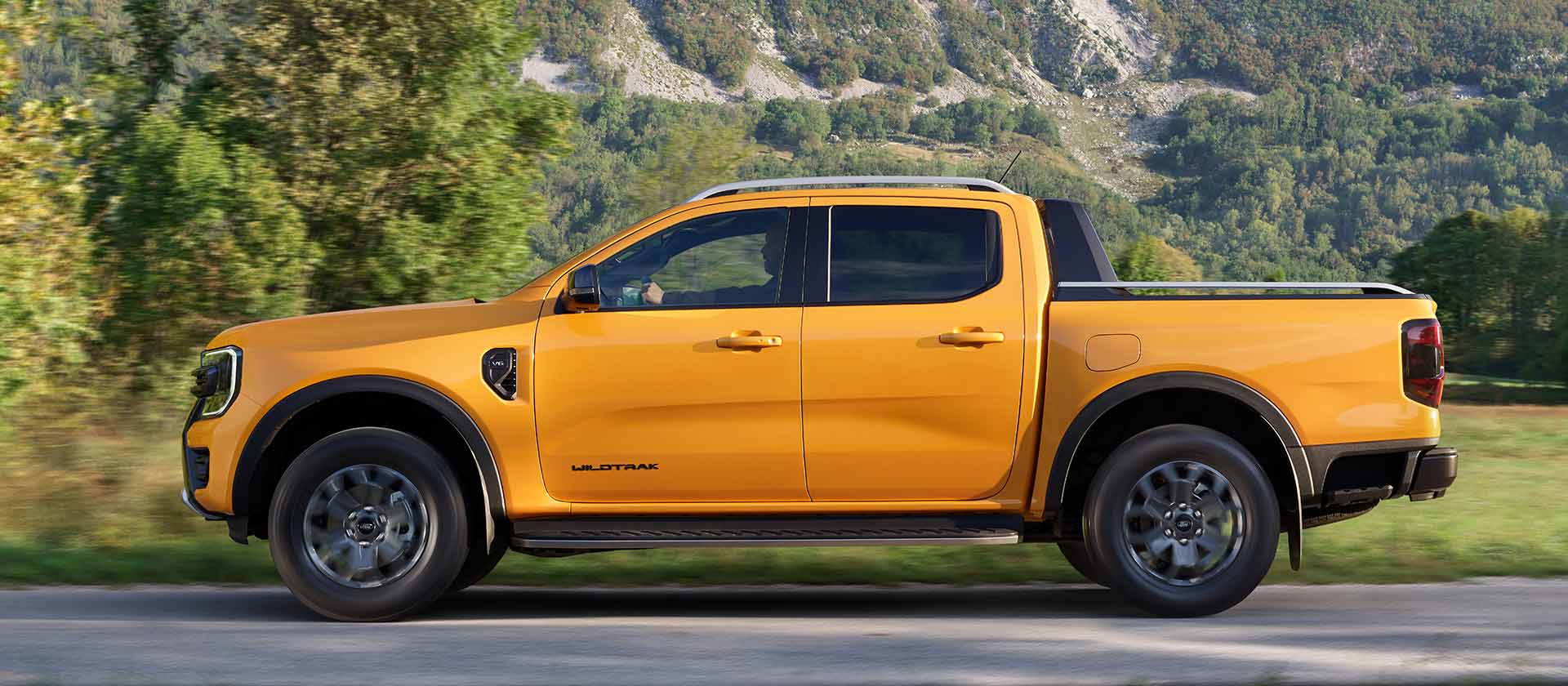 Profil boczny żółtego Forda Ranger Wildtrak z podwójną kabiną zaparkowanego na poboczu drogi w górskiej scenerii.