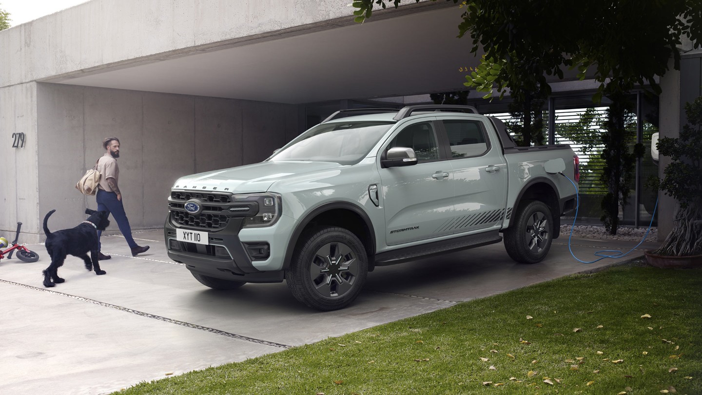Mężczyzna z psem wchodzi do nowoczesnego domu, mijając na zaparkowanego i podłączonego do ładowarki Forda Rangera Stormtrak PHEV