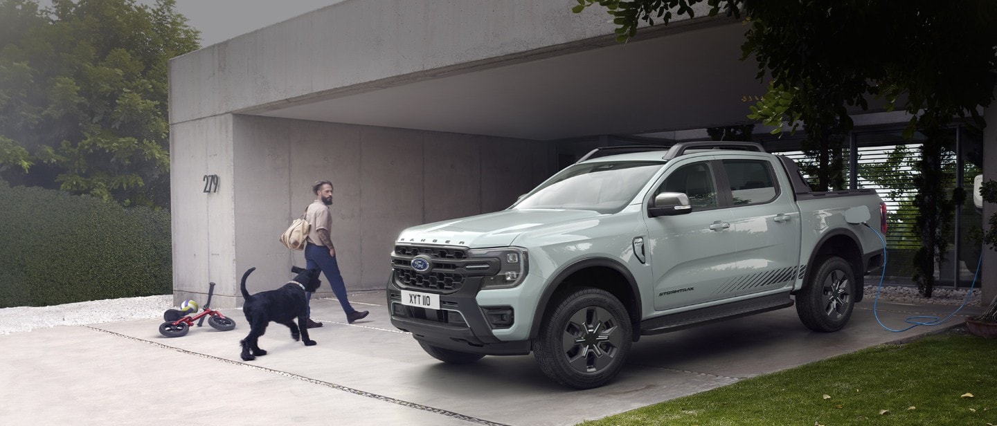 Mężczyzna z psem wchodzi do nowoczesnego domu, mijając na zaparkowanego i podłączonego do ładowarki Forda Rangera Stormtrak PHEV. 