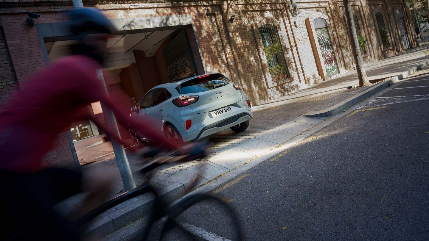 Szary Ford Puma wyjeżdża tyłem z podjazdu, rowerzysta w czerwieni przejeżdża.