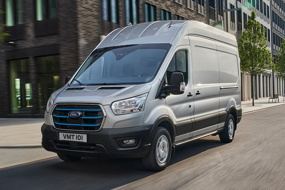 Srebrny Ford Transit jadący przez miejski krajobraz.