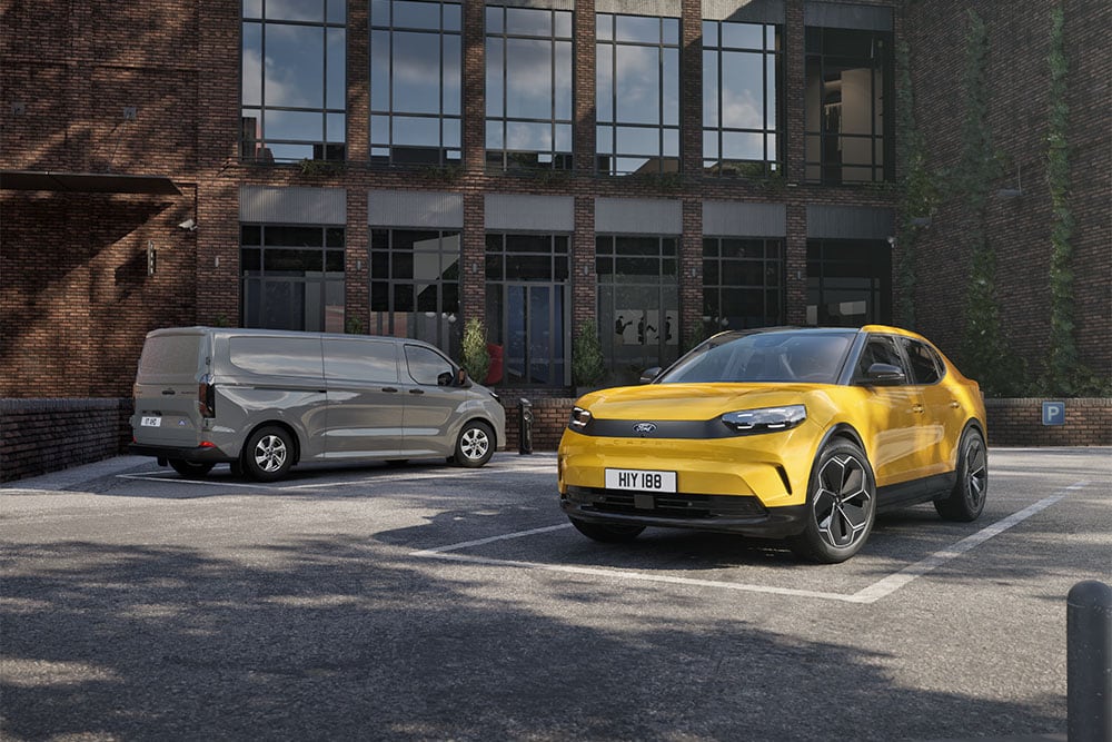  Szara furgonetka Ford i żółty Ford Capri zaparkowane przed nowoczesnym ceglanym budynkiem.