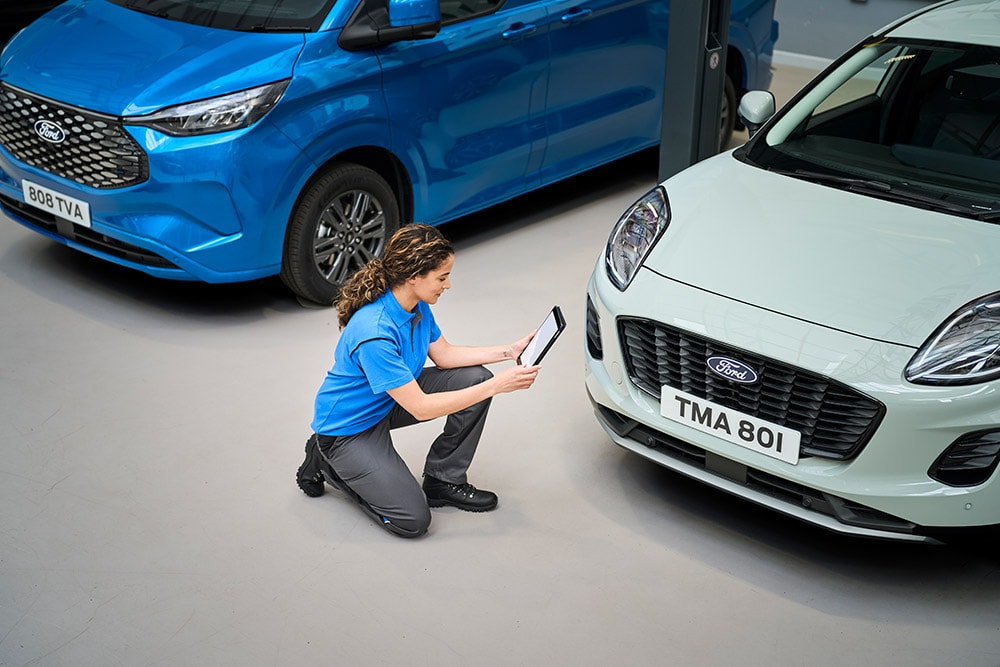 Technik klęczy pomiędzy niebieską furgonetką Ford a białym Fordem Puma i patrzy na tablet.