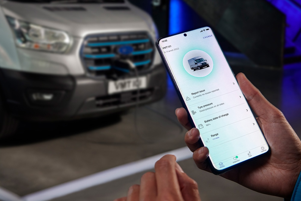 Osoba trzyma smartfon z aplikacją Forda, a w tle ładuje się Ford E-Transit Custom.