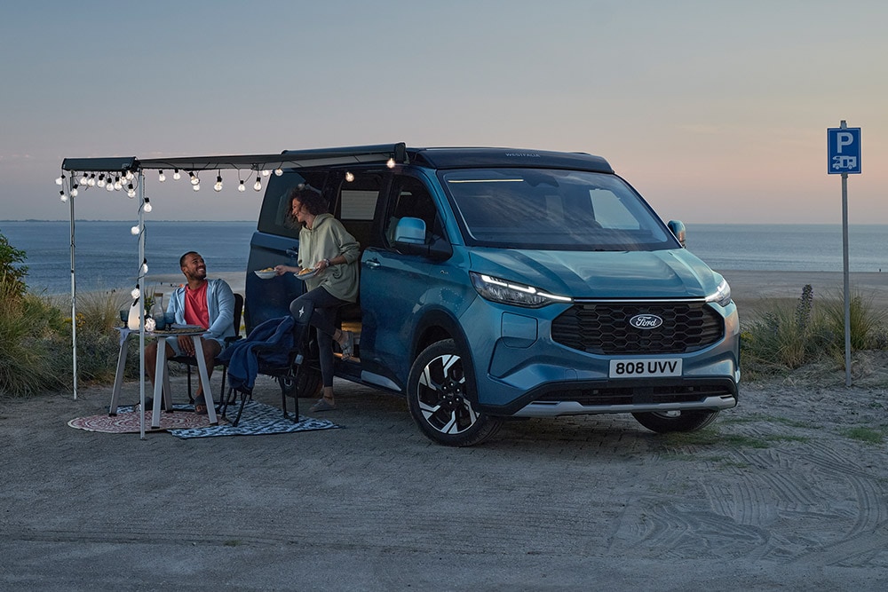  Dwóch kamperów relaksujących się przy niebieskim Fordzie Pro Transit Custom Nugget, w tle parking przy plaży.