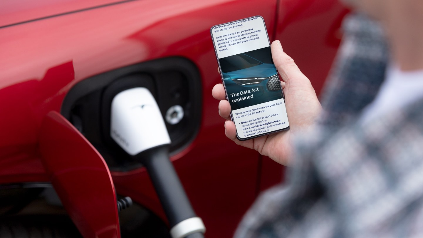 Osoba sprawdzająca smartfon, a w tle ładujący się czerwony samochód elektryczny.
